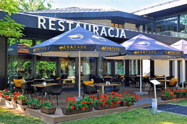 Taras restauracji pod granatowymi parasolami z logo EVA Park, skrzynki z czerwonymi pelargoniami, na budynku napis RESTAURACJA