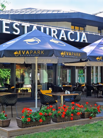 Taras restauracji pod granatowymi parasolami z logo EVA Park, skrzynki z czerwonymi pelargoniami, na budynku napis RESTAURACJA